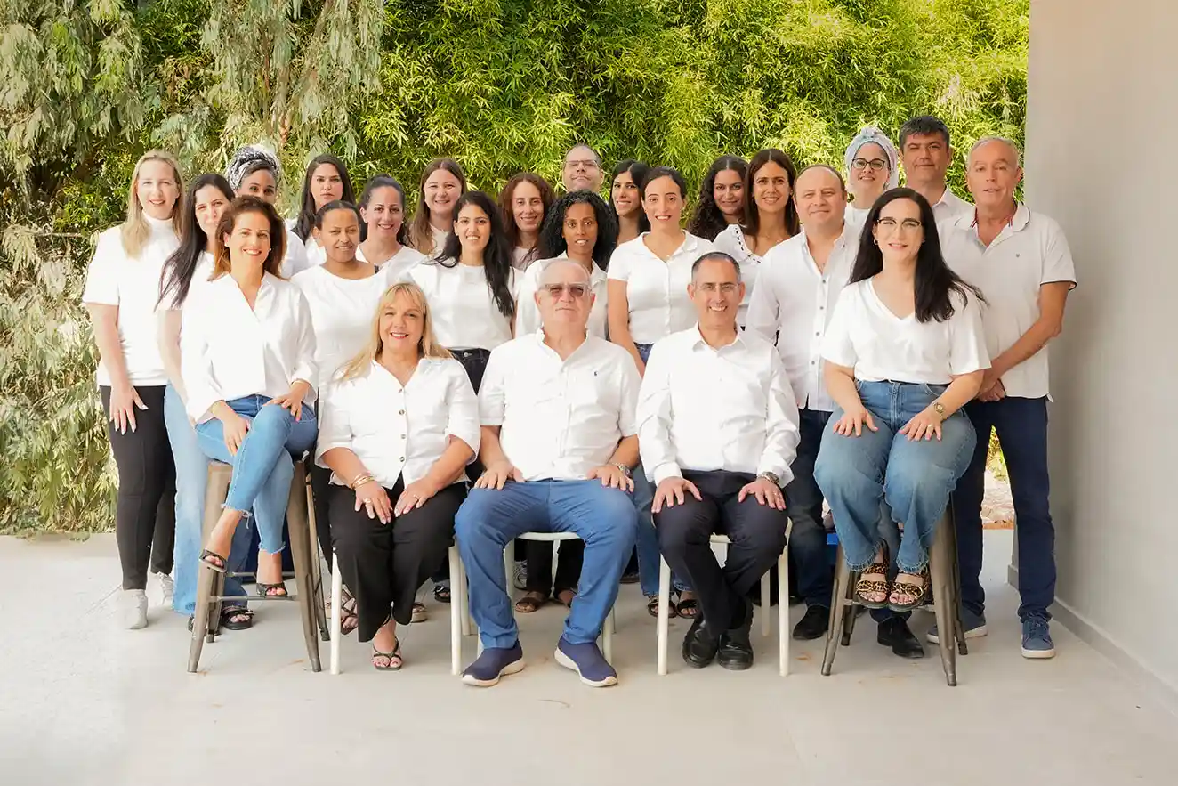 תמונה קבוצתית של צוות אביב - שלוש שורות של כ-24 אנשים, הראשונים ישובים על כיסאות ומאחורים עומדים אנשים נוספים. נראים בתמונה אנשים בגילאים, במגדרים וממגזרים שונים. כולם לבושים חולצה לבנה ומכנס/חצאים כחולים/כהים.